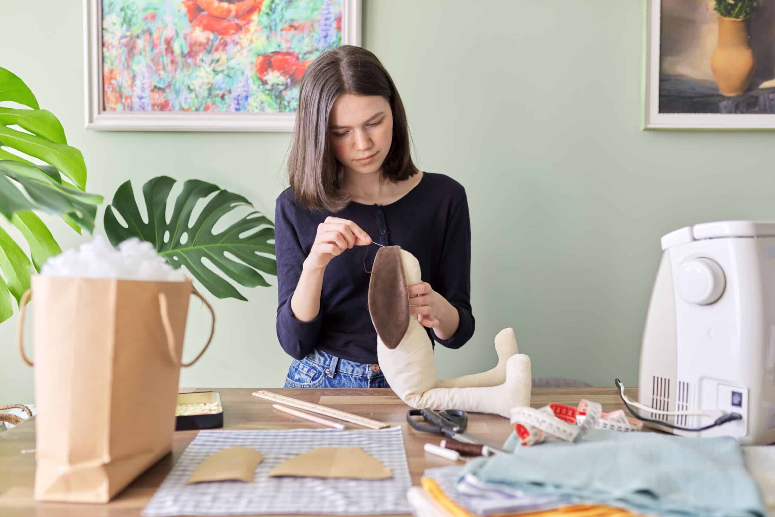 Teen girl sewing toy bunny doll sitting at home at table