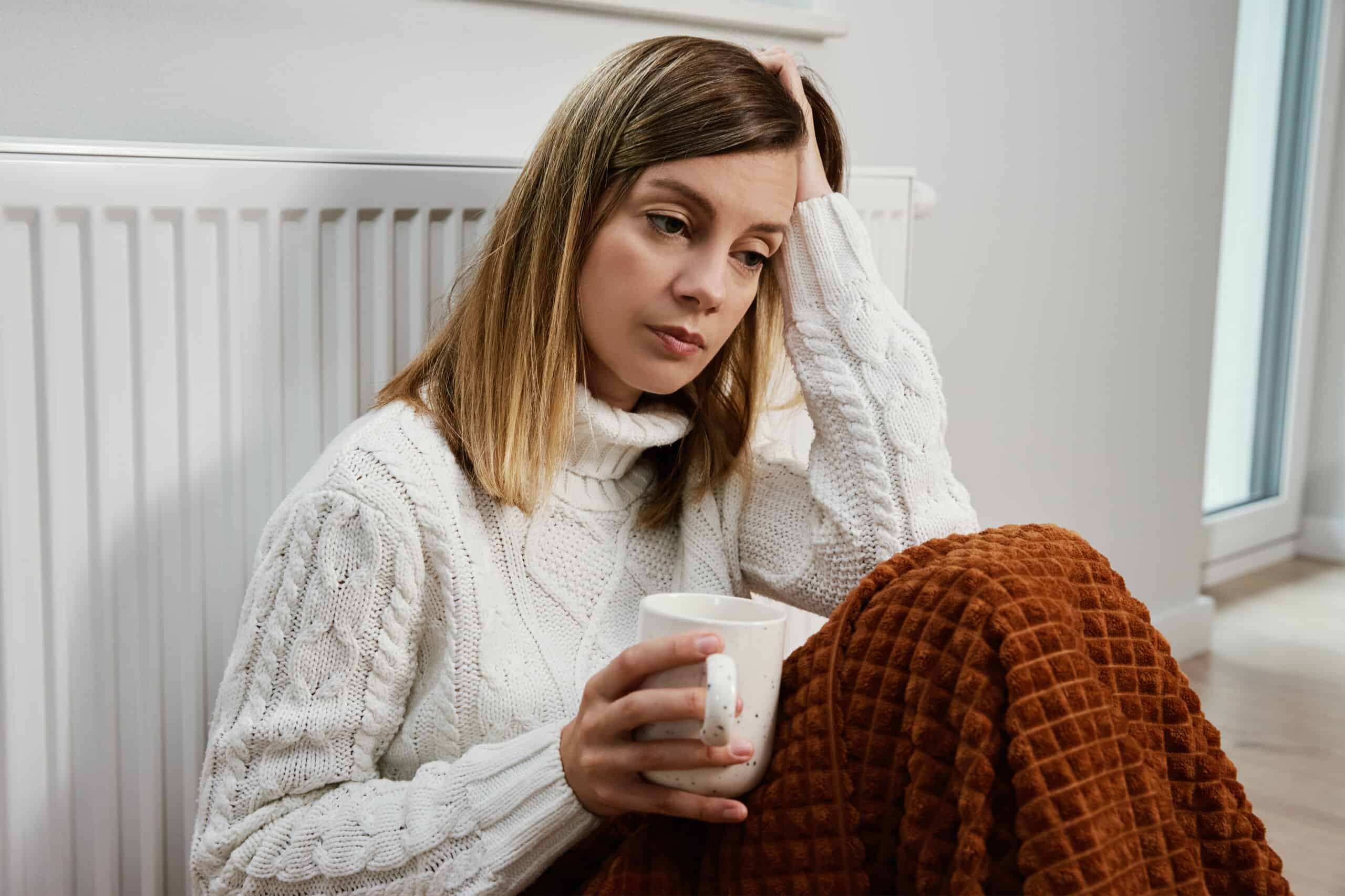 Worried woman sit near heating radiator under blanket with cup of tea