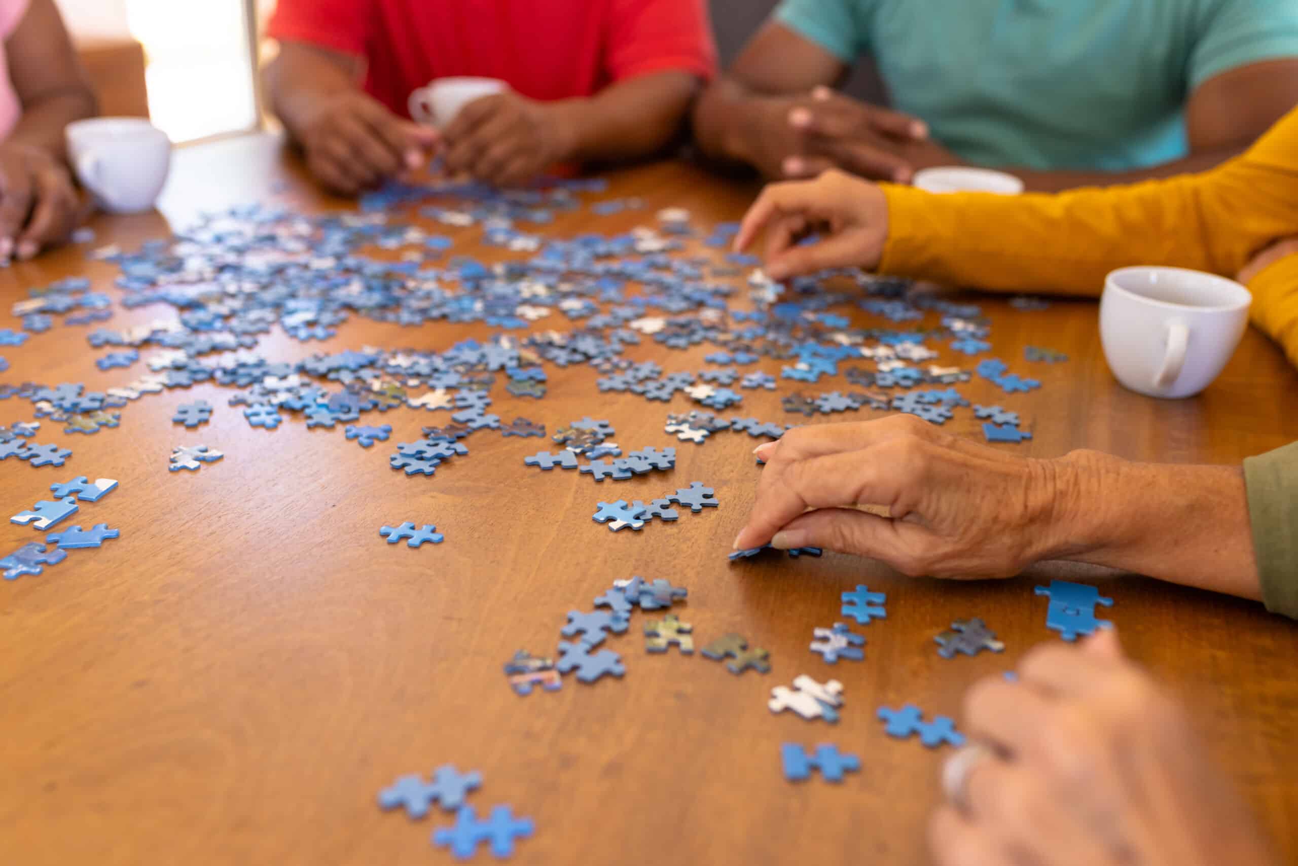 Cropped hands of multiracial seniors arranging jigsaw pieces on wooden table in nursing home. Puzzle, game, confusion, brainstorming, unaltered, togetherness, support, assisted living, retirement.